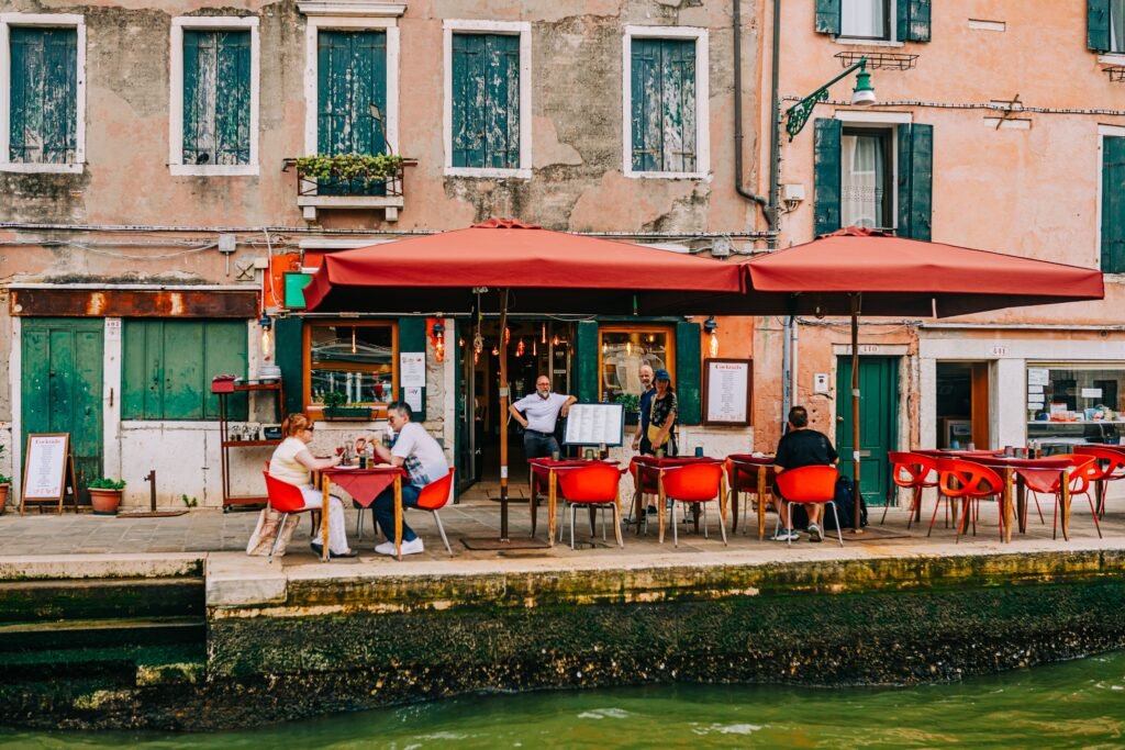 Charming outdoor café by a Venetian canal, featuring vibrant red chairs and patrons enjoying a relaxed day.